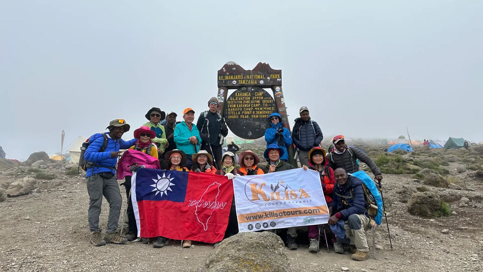 Trekking group holding a Kilisa Tours banner and the flag of Taiwan at the Karanga Camp sign