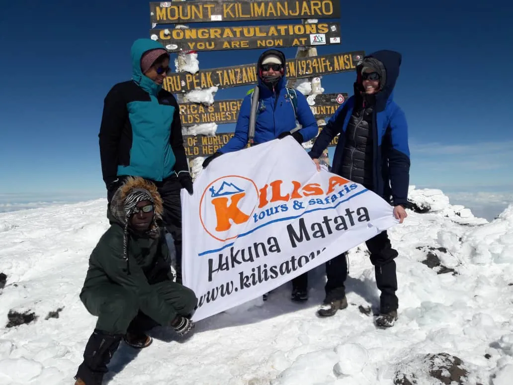 Climbers holding a Kilisa Tours & Safaris banner in the snow at Uhuru Peak, the summit of Mount Kilimanjaro