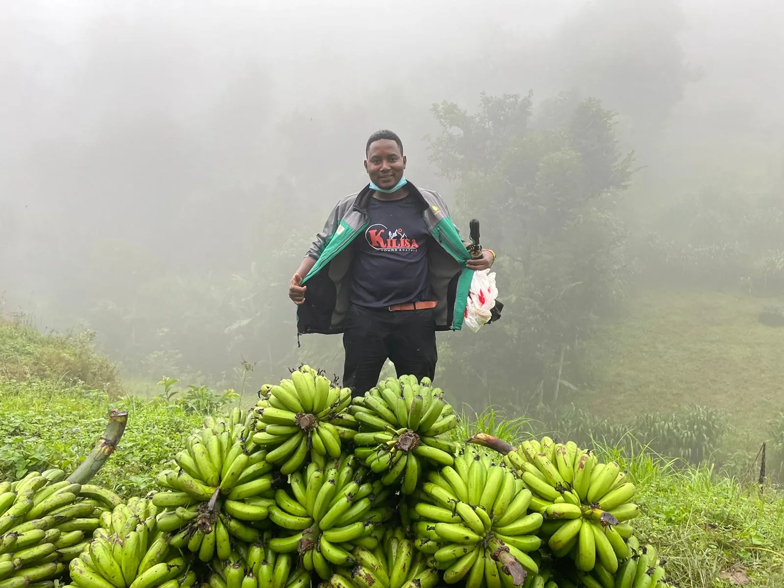 Kilisa Tours guide standing behind a pile of freshly harvested green bananas on a misty village tour in Tanzania
