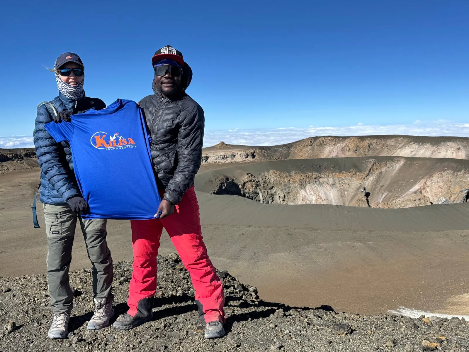Two men holding a blue Kilisa Tours shirt on the rim of Kilimanjaro's summit crater