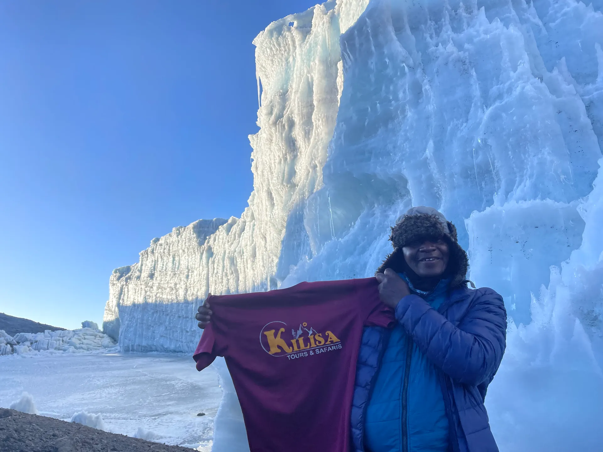 Guide holding a Kilisa Tours & Safaris shirt in front of a glacier ice wall near the summit of Kilimanjaro