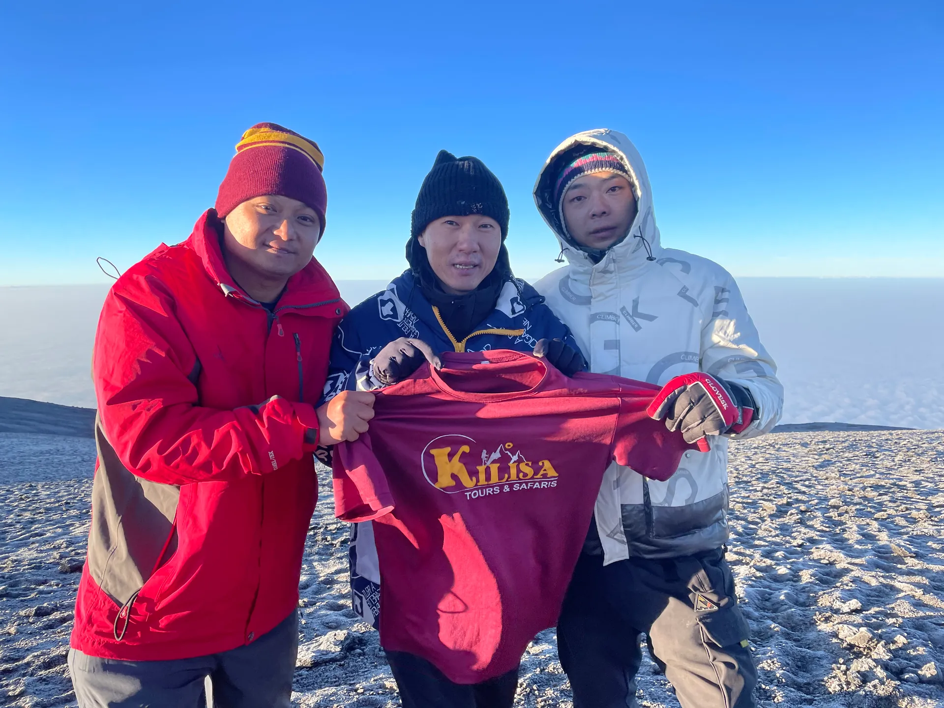 Three climbers holding a Kilisa Tours shirt on the frosty summit of Kilimanjaro at sunrise